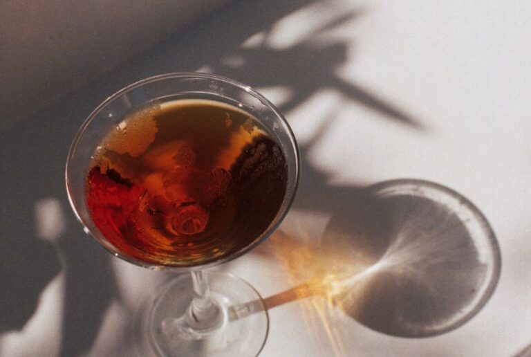 A vibrant red-colored cocktail in a martini glass on a white stair with sunlight reflection and leaf shadows
