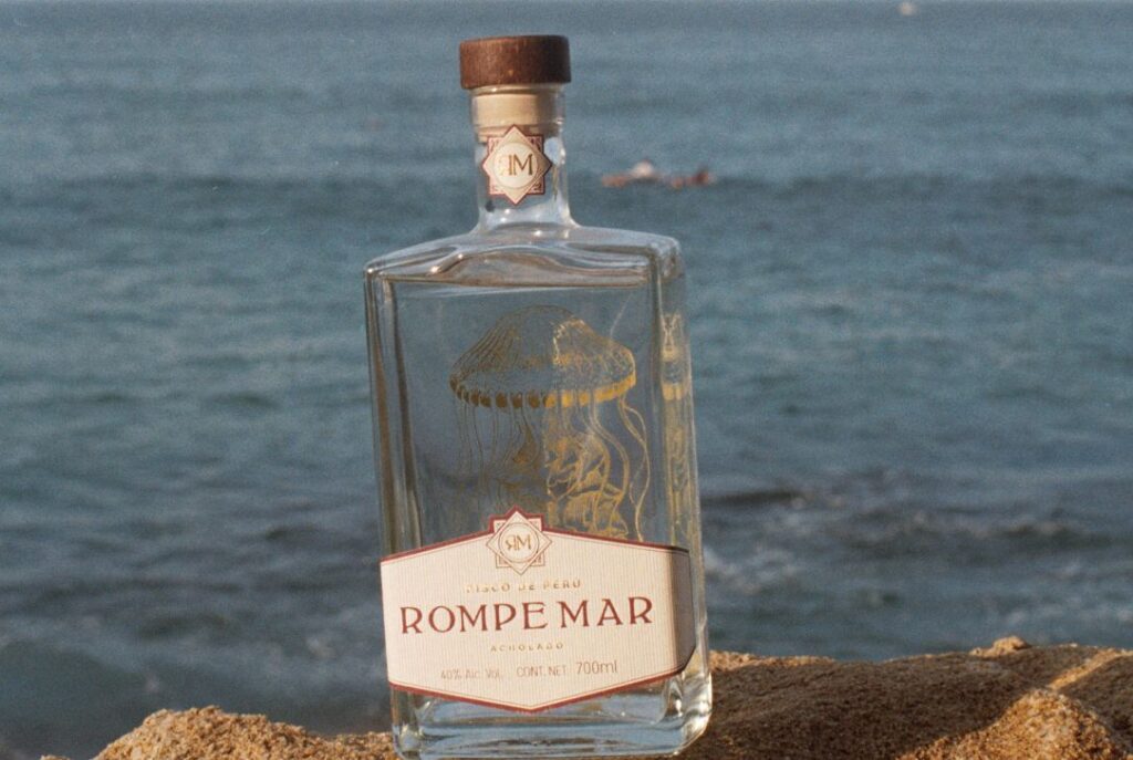 Rompe Mar Pisco bottle on a rock with golden jellyfish symbol and ocean in the background, with sunlight shining on the bottle