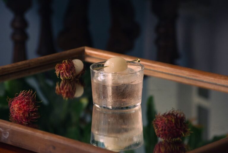 Pisco Lychee, with a light rose colour, garnished with lychee, surrounded by lychees with a bright pink/red outside, on a squared mirror with a brown frame